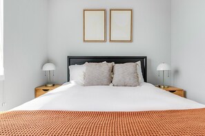 Een net opgemaakt bed met een witte dekbedovertrek, twee kussens en een geribbeld oranje dekbed. Er staan twee nachtkastjes met lampen bij, en de muren zijn effen met twee ingelijste foto’s.