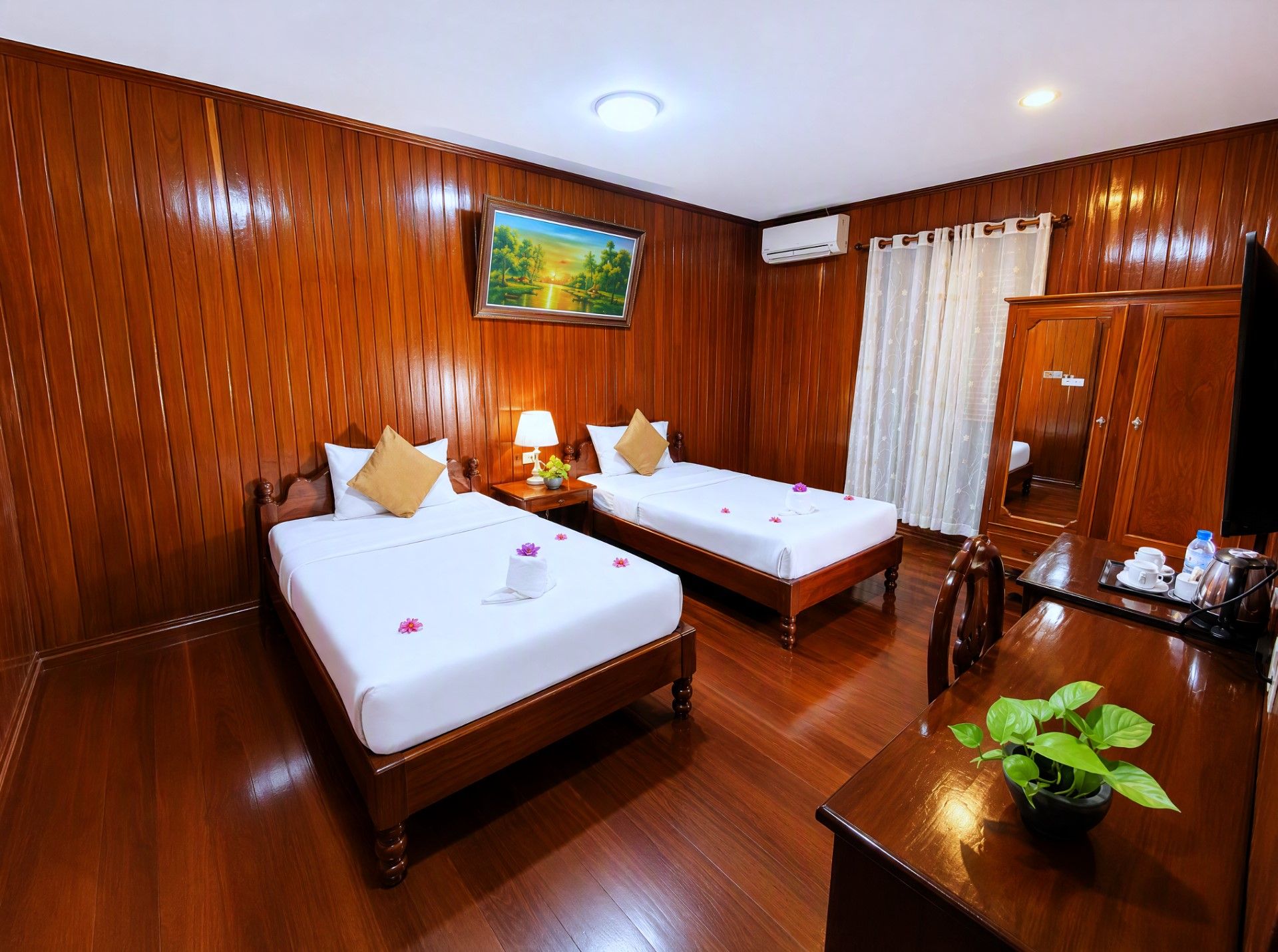 Une chambre d’hôtel avec deux lits en bois, un tableau au mur, un appareil de climatisation, un bureau avec une plante et un miroir.