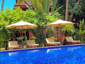 Un espace aménagé au bord de la piscine, avec des chaises longues et des parasols, entouré d’une végétation luxuriante et bordé d’un bâtiment au toit rouge.