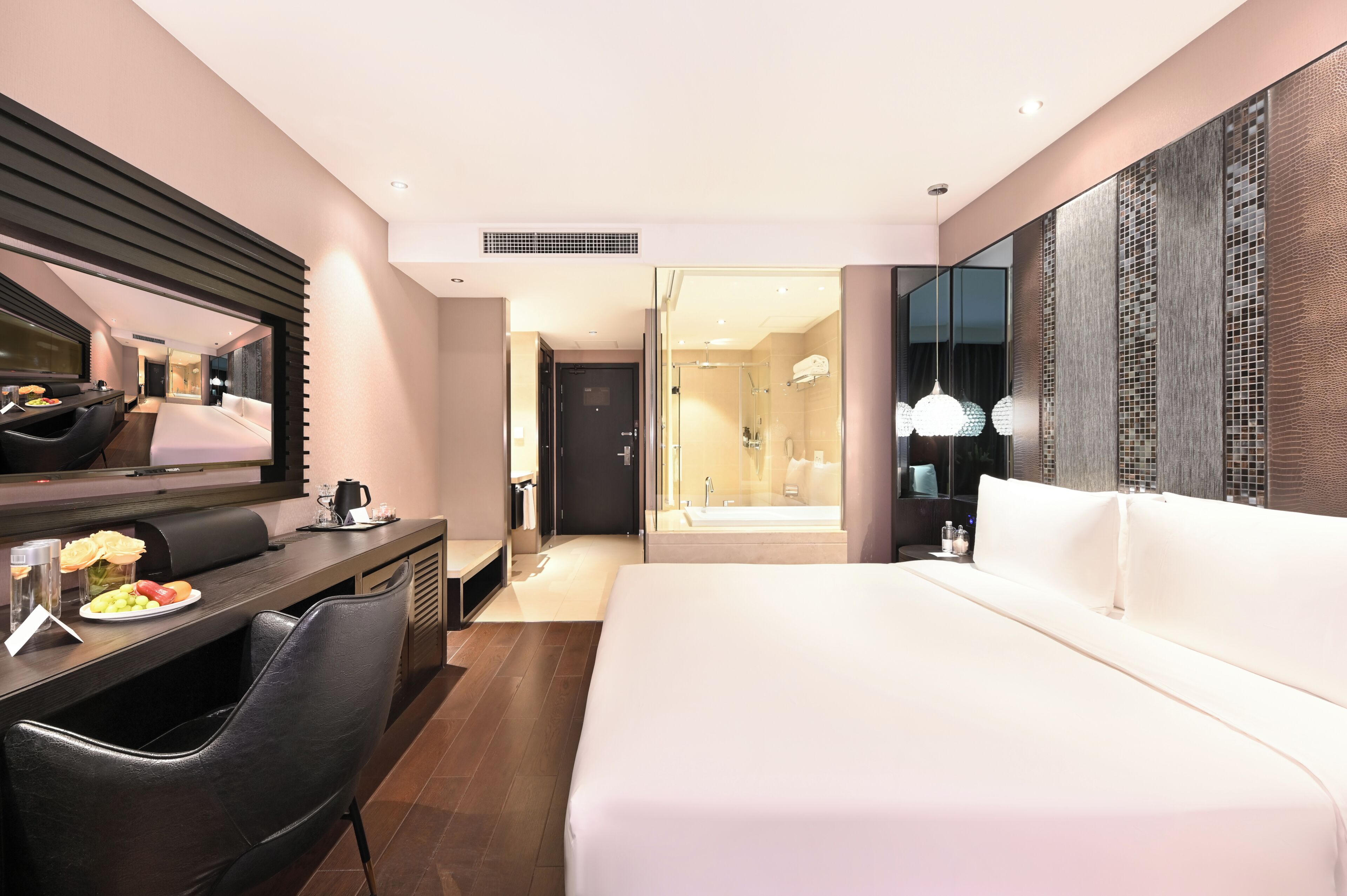 Habitación de hotel moderna con una cama grande, un escritorio con silla, un espejo y un baño con ducha y lavamanos.