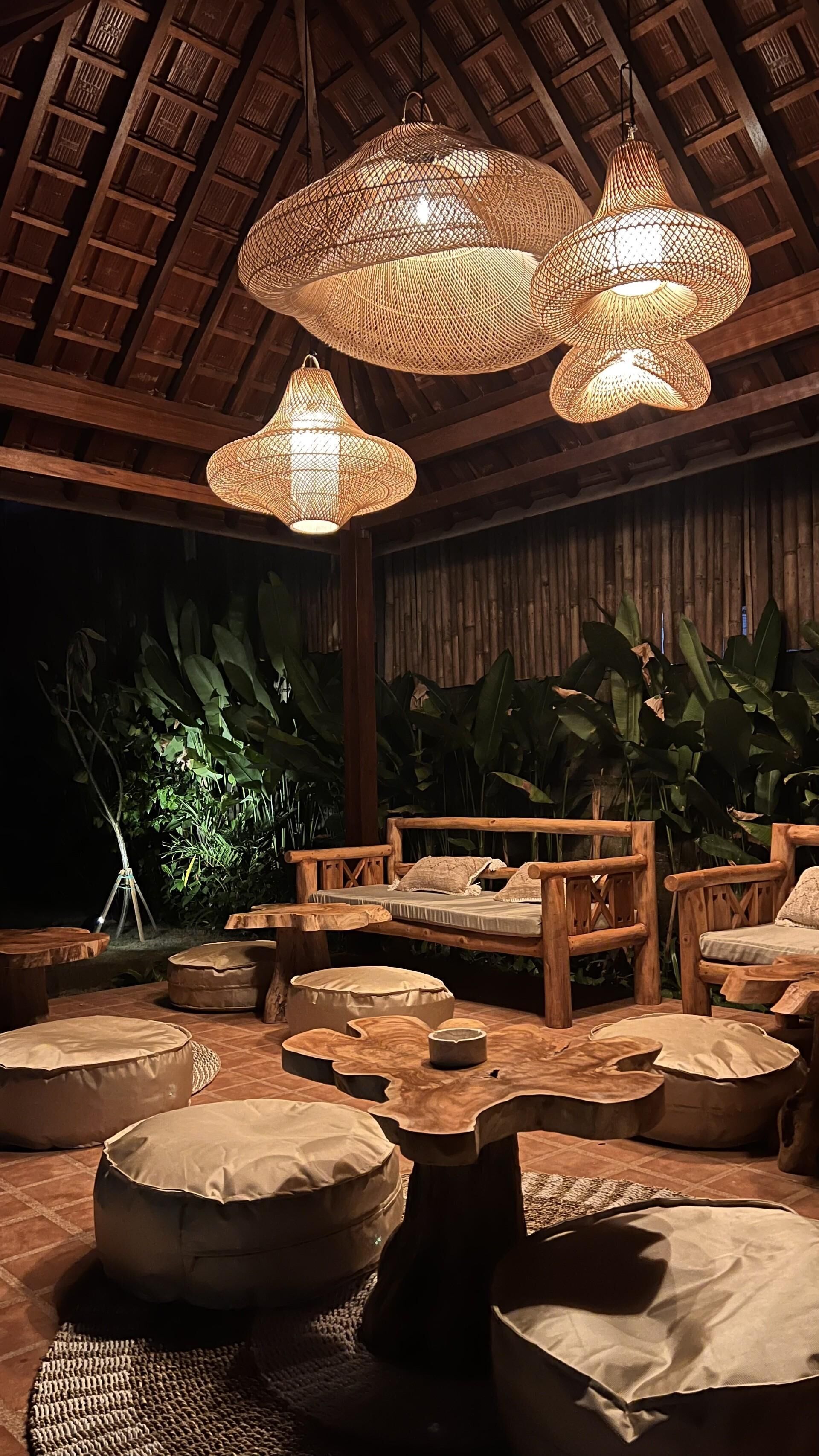 A cozy outdoor seating area with woven light fixtures, wooden furniture, and lush greenery.