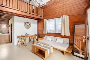 Une pièce spacieuse avec un plafond en bois, un coin repas comprenant une table et des chaises en bois, un canapé avec des coussins et une grande fenêtre avec des rideaux.