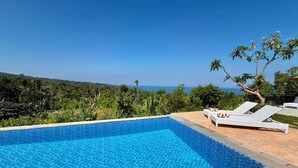 Une piscine d’un bleu éclatant, dotée de chaises longues, entourée d’une végétation luxuriante et offrant une vue sur l’océan.