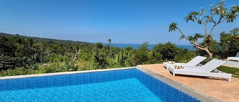 A clear blue swimming pool with lounge chairs, surrounded by lush greenery and a view of the ocean.