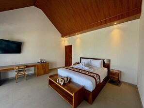 A modern bedroom with a wooden ceiling, a large bed with white bedding, a wooden desk, a chair, and a TV mounted on the wall. - Atta Residence (Karangasem)
