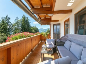 Un balcón de madera con un sofá y sillones, una mesa con una botella y vasos, y vistas a árboles y montañas.
