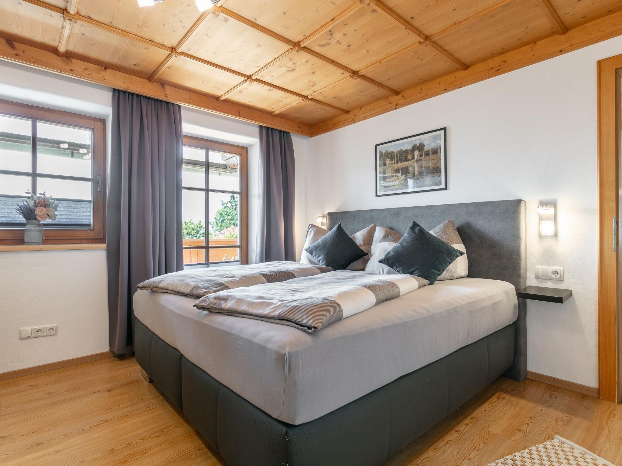 Une chambre avec un plafond en bois, un lit avec une literie grise, une fenêtre avec des rideaux et un luminaire fixé au mur.