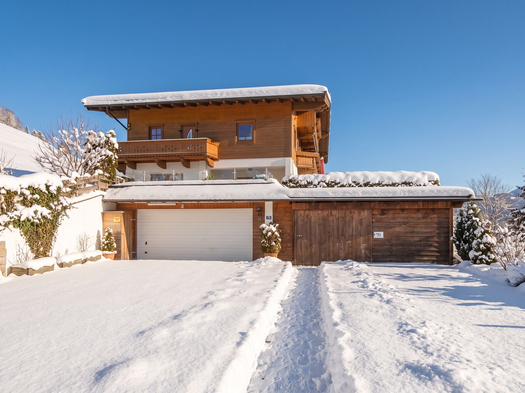 A two-story wooden house with a garage, surrounded by snow-covered ground and trees.