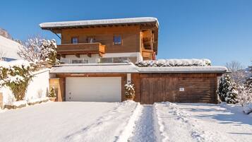 A two-story wooden house with a garage, surrounded by snow-covered ground and trees.
