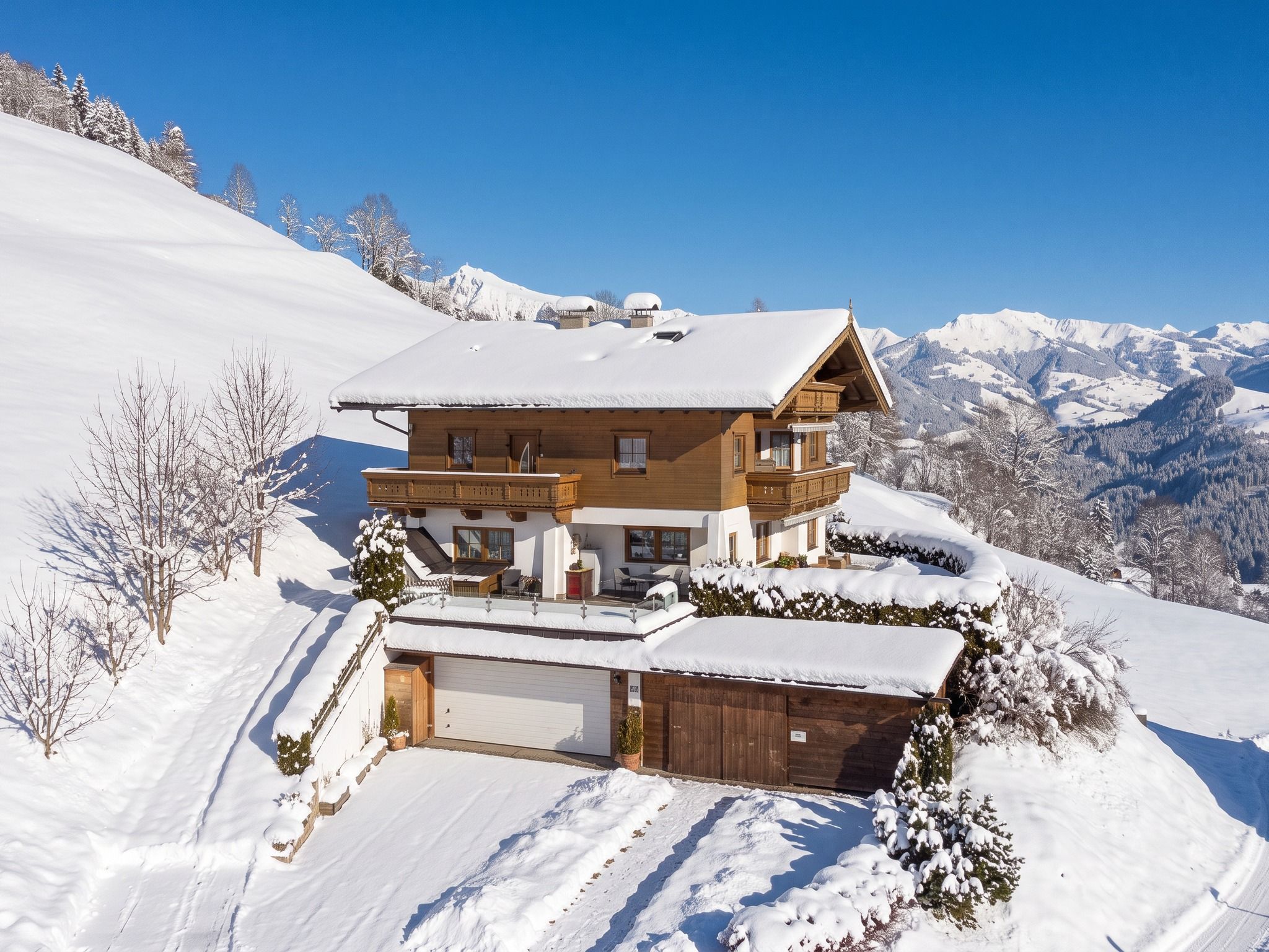 Un chalet en bois à deux étages, dont le toit est recouvert de neige, situé sur une pente enneigée avec une chaîne de montagnes en arrière-plan.