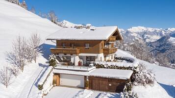 Un chalet en bois à deux étages, dont le toit est recouvert de neige, situé sur une pente enneigée avec une chaîne de montagnes en arrière-plan.