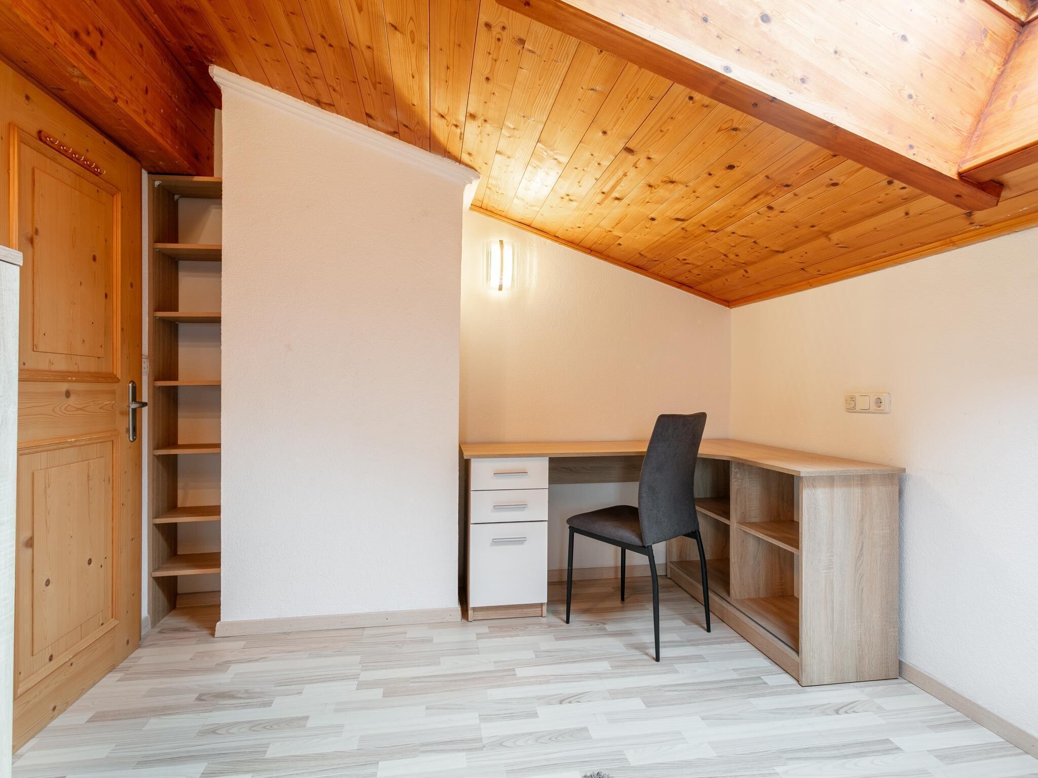 A room with a wooden ceiling, a desk with a chair, a wardrobe, and a built-in shelving unit.