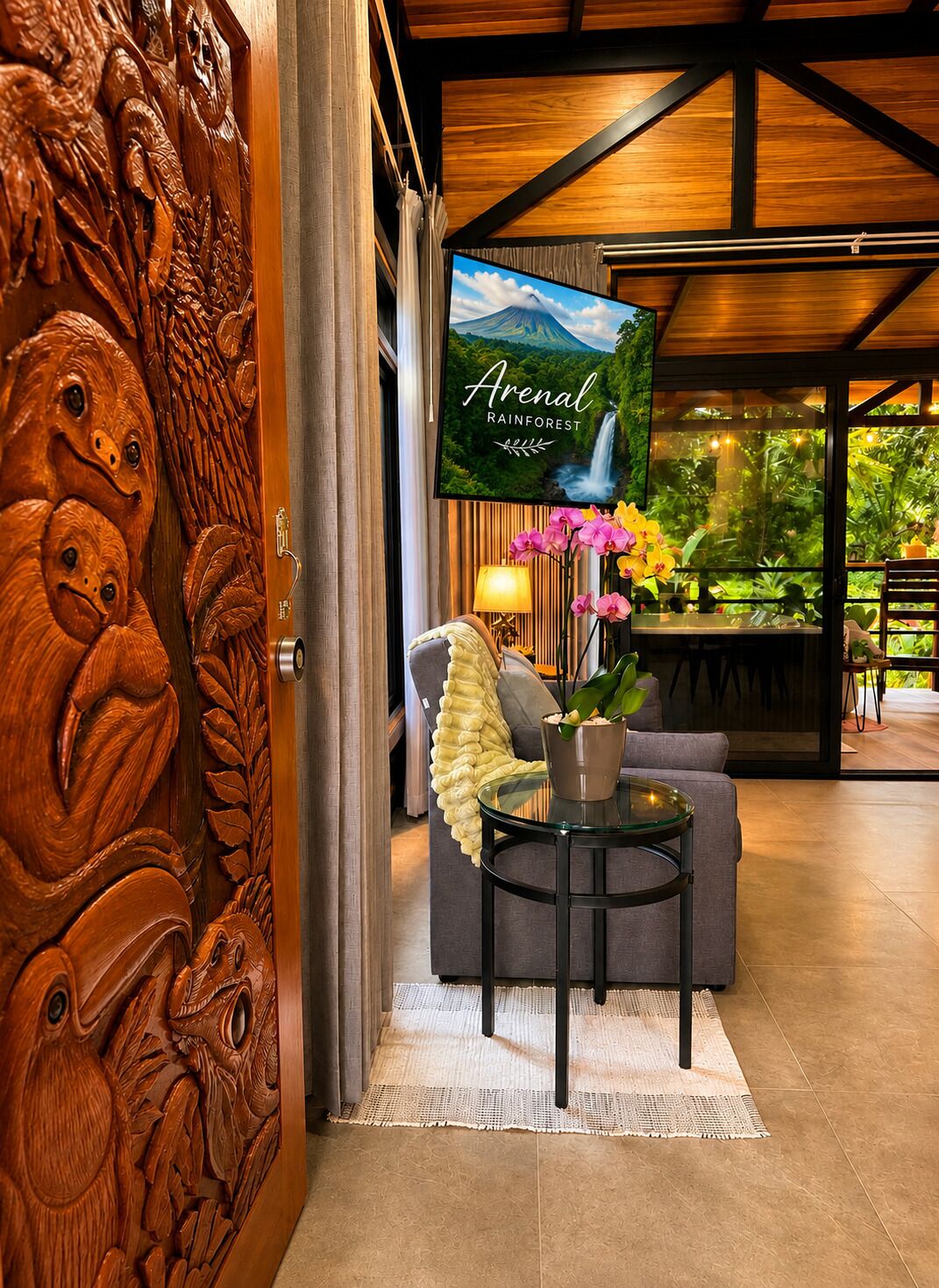 Una habitación con una puerta de madera tallada, un televisor que muestra una escena de selva tropical, un sofá, una mesa con una planta y vistas a la vegetación exterior.