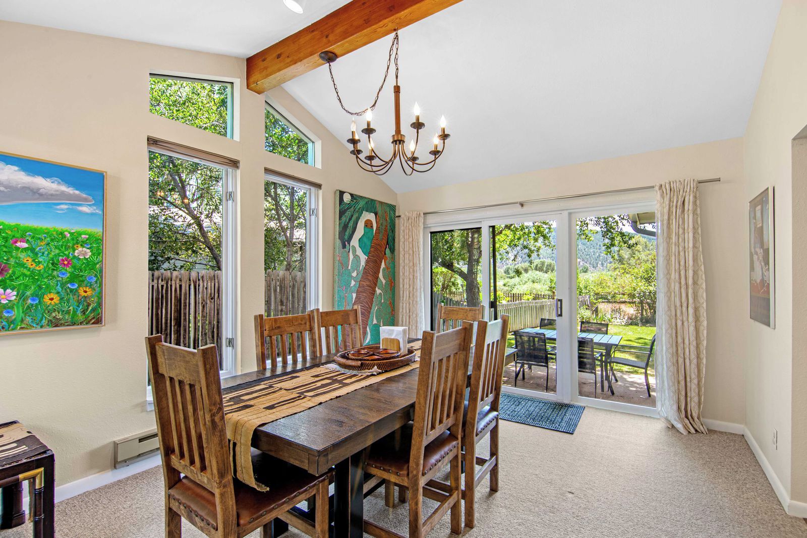 A dining area with a wooden table and chairs, a chandelier, and a view of the garden through large windows.