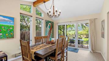 A dining area with a wooden table and chairs, a chandelier, and a view of the garden through large windows.