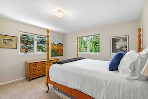A bedroom with a four-poster bed, a wooden dresser, and two windows with artwork.