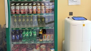 Een automaat gevuld met drankjes en snacks, een wandairconditioner en een ingelijst kunstwerk.