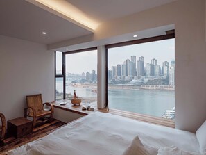 A hotel room with a large bed, a wooden desk, a chair, and a view of a city skyline and water. - Boys2men (Chongqing)