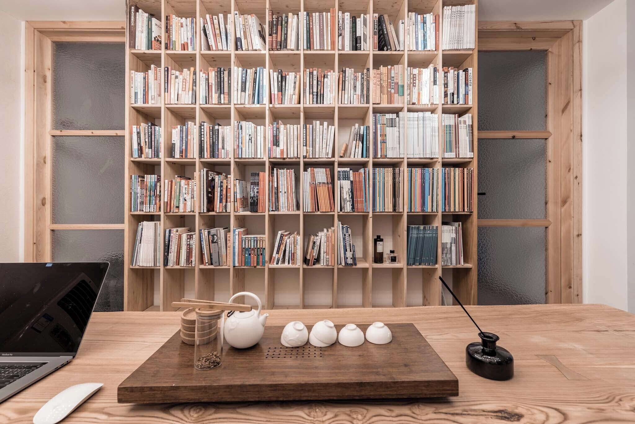 Una mesa de madera con una computadora portátil, un hervidor de agua, tazas y un pequeño objeto negro. Al fondo, una estantería llena de libros.