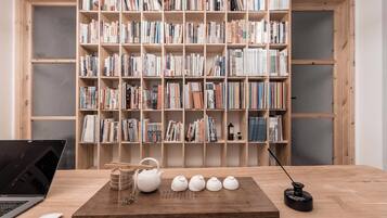 Una mesa de madera con una computadora portátil, un hervidor de agua, tazas y un pequeño objeto negro. Al fondo, una estantería llena de libros.