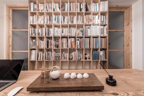 Una mesa de madera con una computadora portátil, un hervidor de agua, tazas y un pequeño objeto negro. Al fondo, una estantería llena de libros.