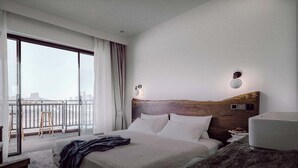 Quarto de hotel com uma cama grande, cabeceira de madeira, mesa de cabeceira, abajur e vista da cidade por uma varanda.