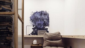Um quarto com estante, um quadro, uma pequena mesa com um jarro branco e um banco acolchoado.