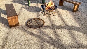 Hiekkapohjainen ulkoalue, jossa on nuotiopaikka, kaksi penkkiä ja metallinen grilli.