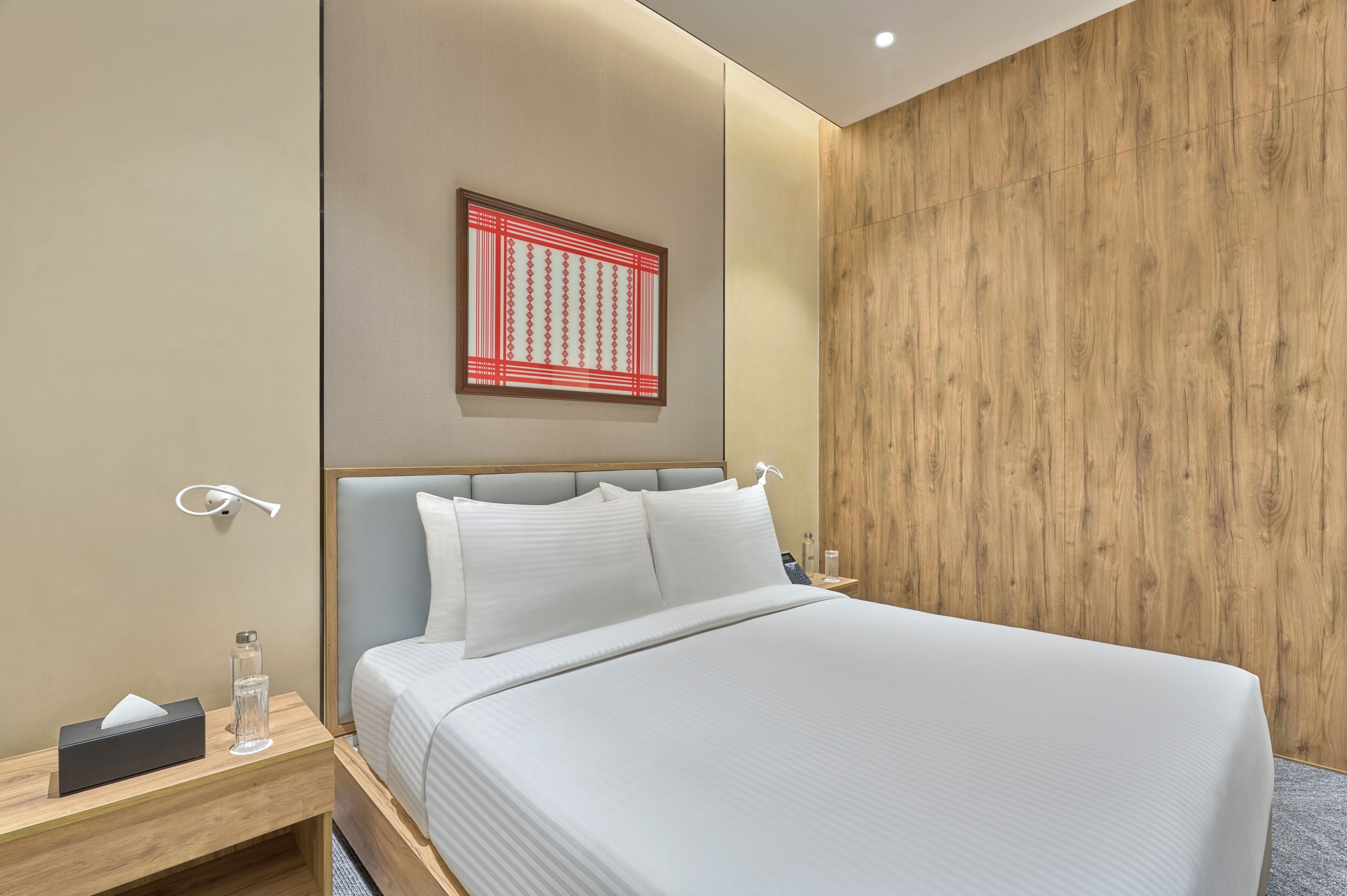 Una habitación de hotel moderna con una cama grande, un panel decorativo de madera, una obra de arte colgada en la pared y una mesita de noche con una botella y una caja de pañuelos.