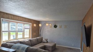 Una sala de estar con sofá modular, televisor de pantalla plana en la pared y ventana con persianas.