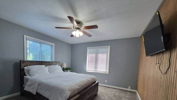 Un dormitorio con cama, ventilador de techo, dos ventanas con persianas y televisor montado en la pared.