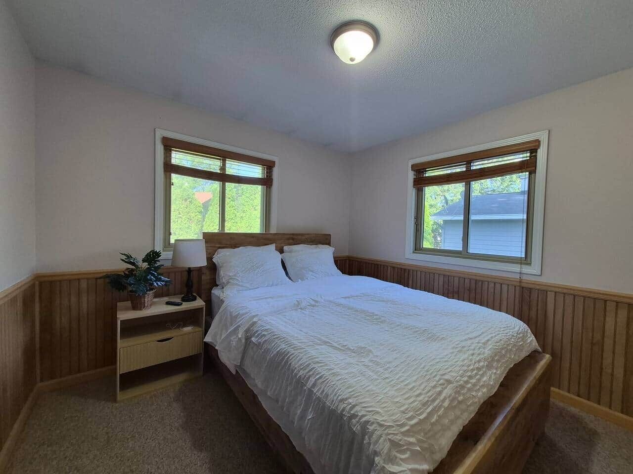 A bedroom with a bed, nightstand, lamp, and windows.