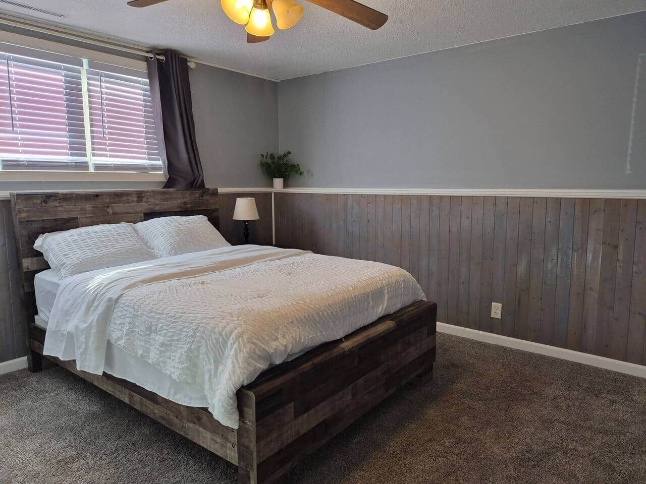 A bedroom with a wooden bed, a ceiling fan, a window with blinds, and a plant.