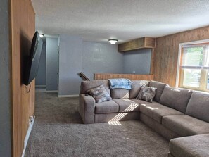 Una sala de estar con un sofá modular, un televisor de pantalla plana montado en la pared y una ventana con persianas.