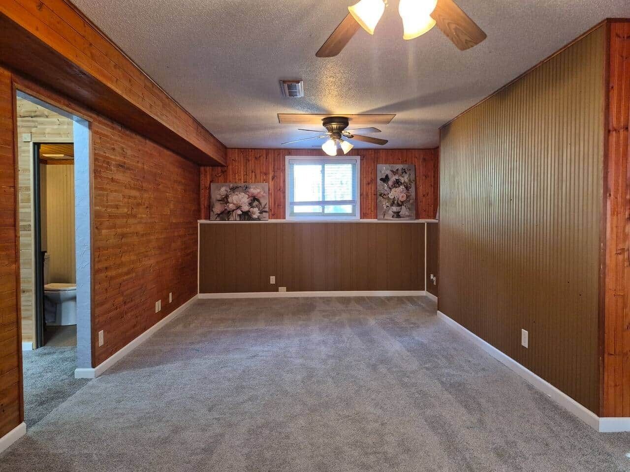 Een kamer met houten wanden, een plafondventilator, een raam en een deur naar een andere kamer.