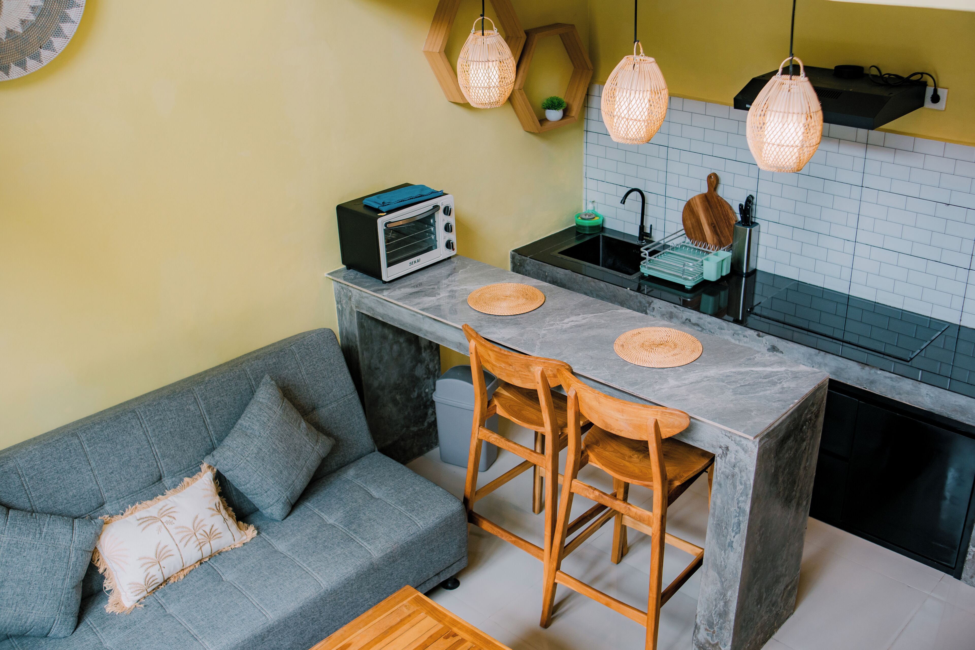 A compact kitchen with a built-in oven, a dining table with chairs, and a grey sofa with a cushion.