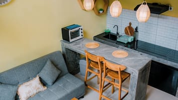 A compact kitchen with a built-in oven, a dining table with chairs, and a grey sofa with a cushion.