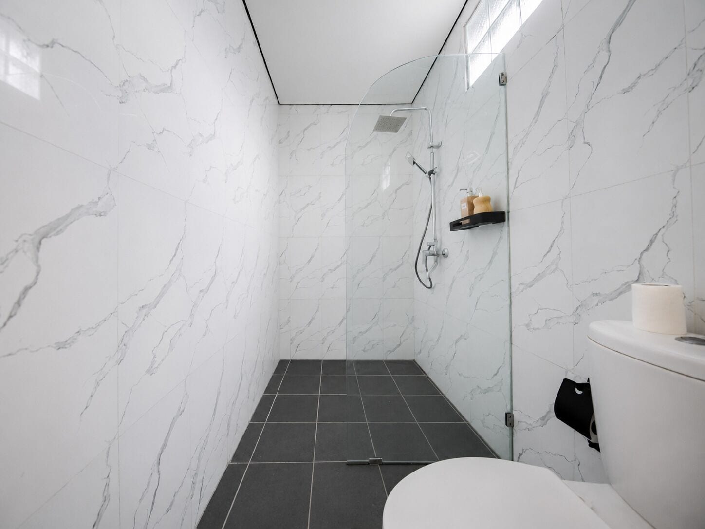 A modern bathroom with white marble walls, a glass shower enclosure, a black tiled floor, and a white toilet.
