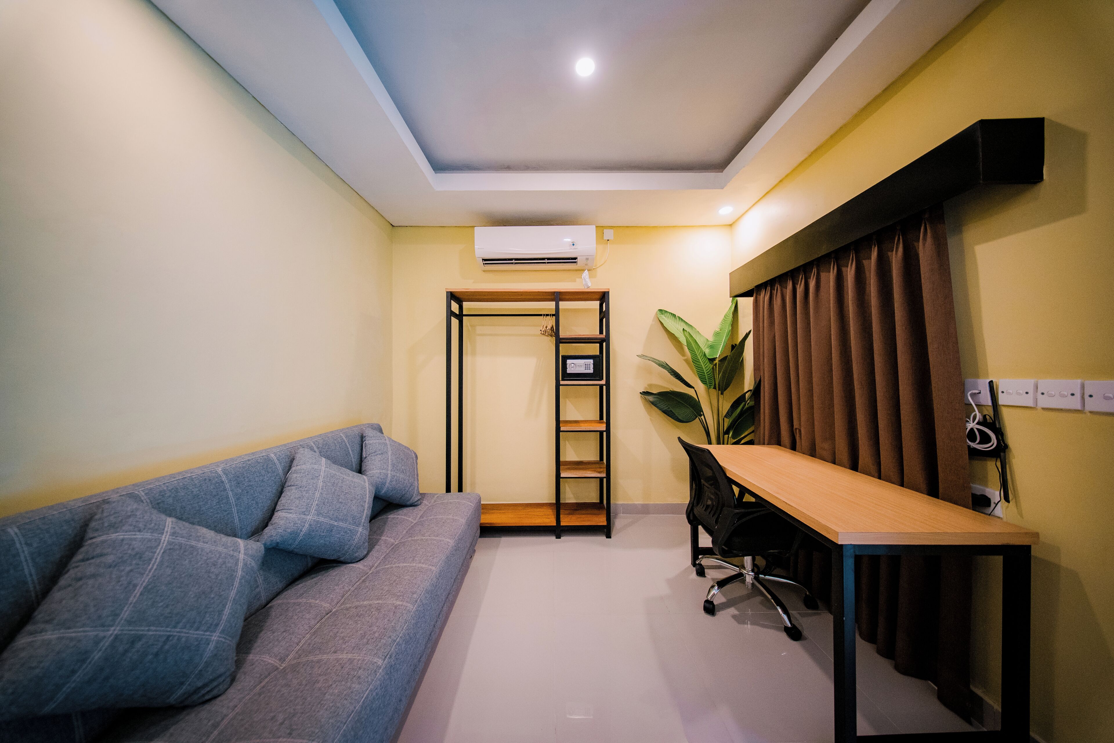 A room with a sofa, a desk, a chair, an air conditioning unit, a plant, and a door.