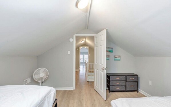 A bedroom with two beds, a dresser, a fan, and a door leading to another room.
