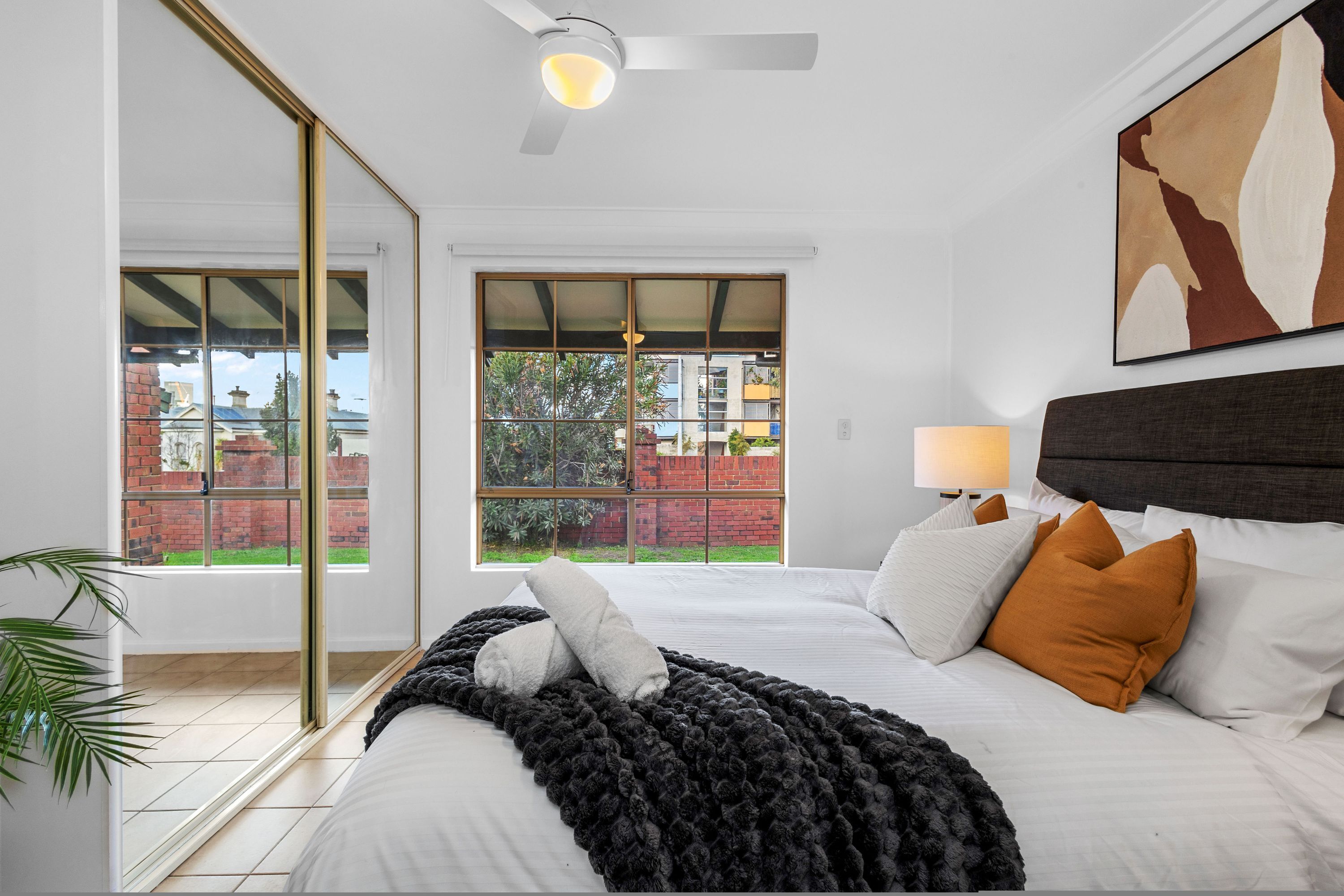 Une chambre avec un grand lit, un ventilateur de plafond, une fenêtre donnant sur un mur de briques et de la verdure, et un tableau accroché au mur.