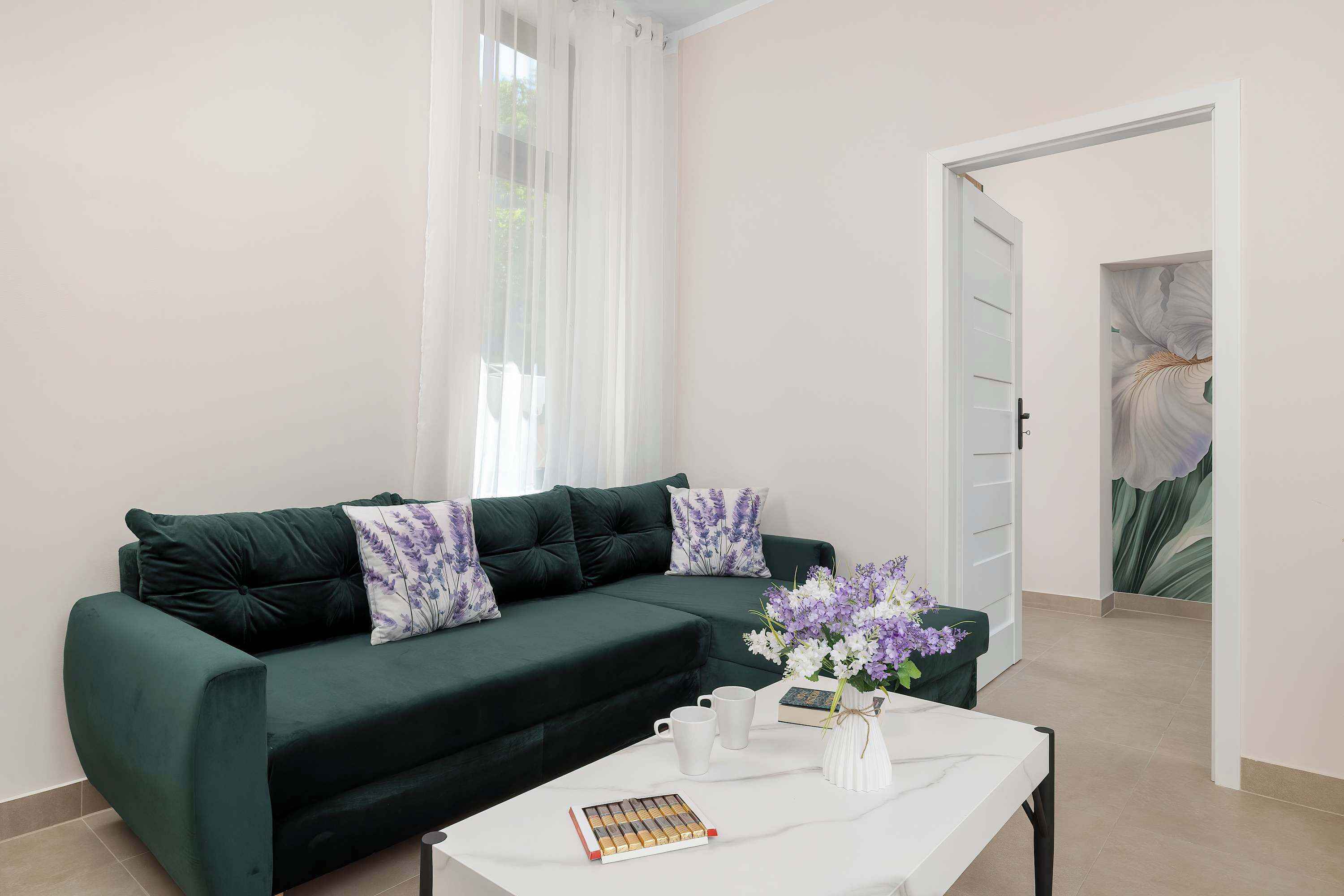 A living room with a dark green sofa, two purple floral cushions, a white coffee table with a book and a vase of flowers.
