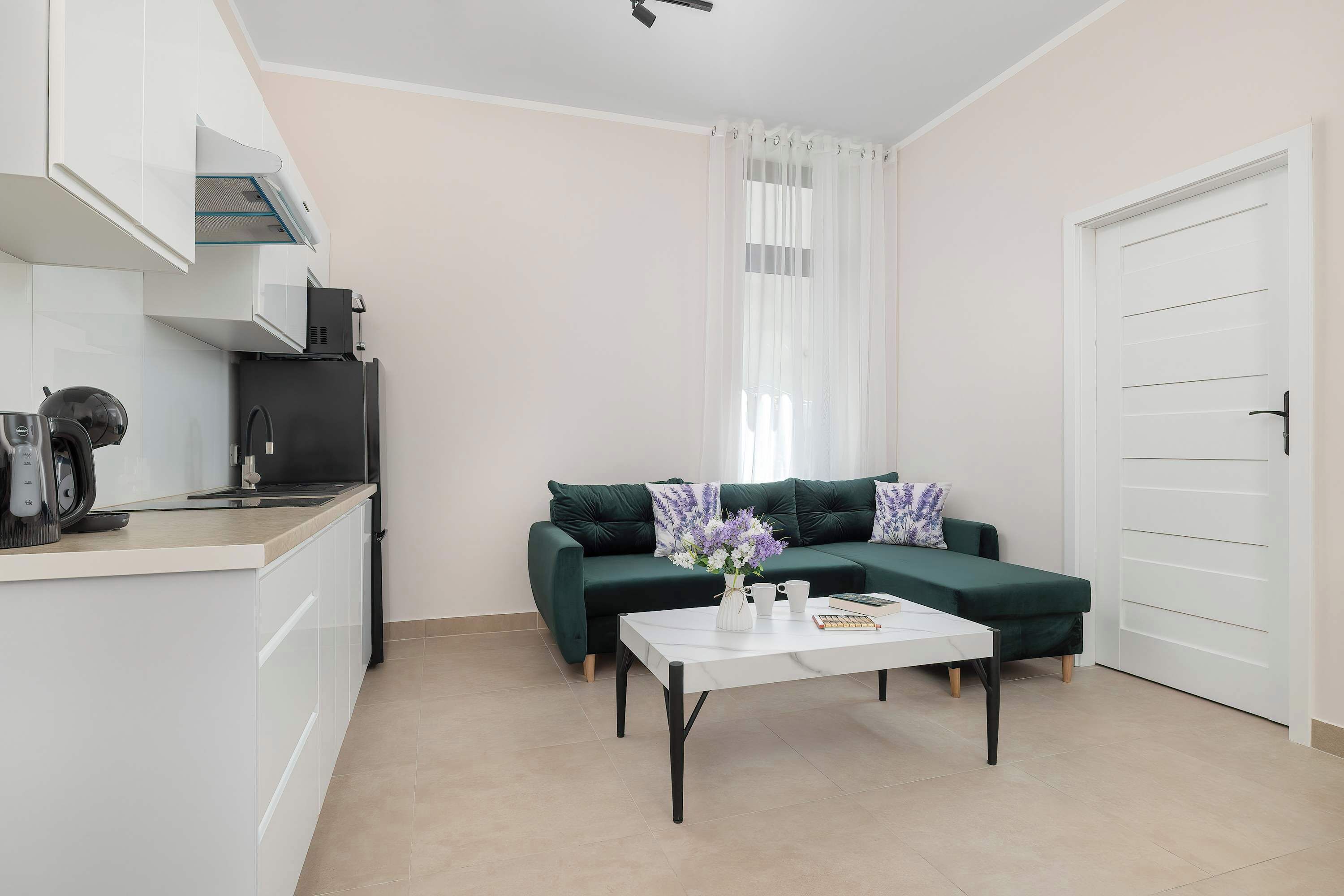 Een moderne keuken met een ingebouwde oven, een koffieapparaat en een koelkast. Er is een eethoek met een tafel en stoelen, en een groene bank met decoratieve kussens.