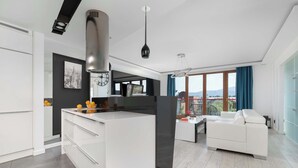 A modern kitchen with a central island, a dining area with a white sofa, and a view of the city through large windows. - Stylish Apartment with a Balcony | Close to City Center (Bielsko-Biała)