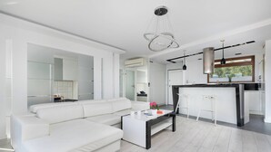 A modern living room with a white sofa, a coffee table, and a kitchen area with a bar and a window. - Stylish Apartment with a Balcony | Close to City Center (Bielsko-Biała)