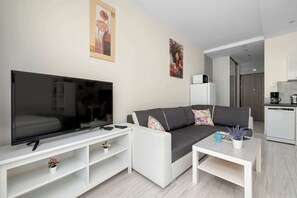Ein modernes Wohnzimmer mit Flachbildfernseher, einer Couch, einem Couchtisch und einer Küchenzeile mit Kühlschrank und Kaffeemaschine.