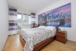 A bedroom with a bed, a nightstand, a lamp, and a cityscape painting on the wall. - Spacious Apartment with Balcony | Workspace | Close to the Center (Warszawa)