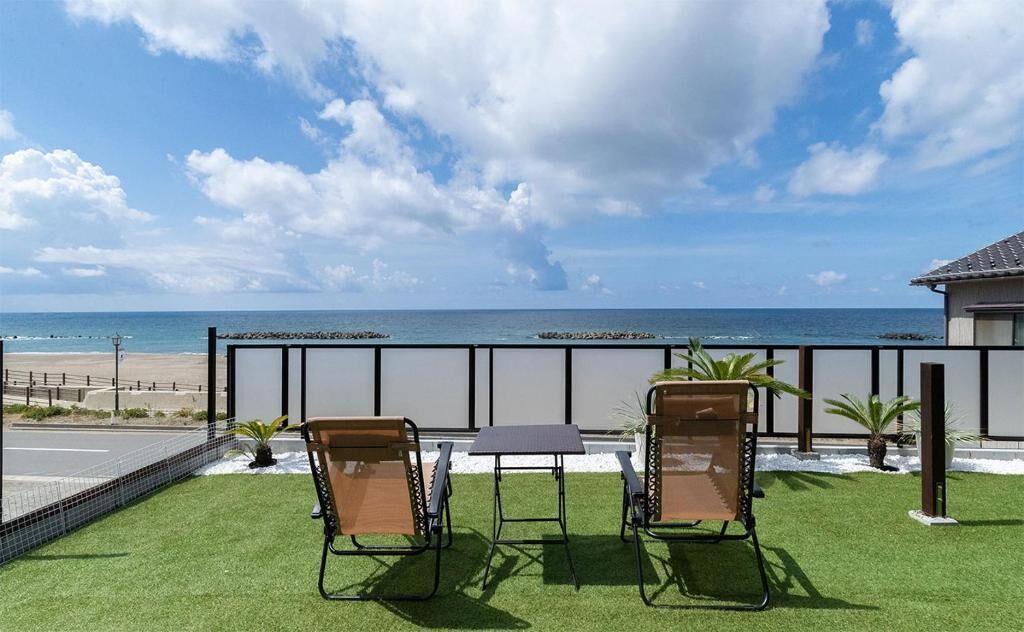 A balcony with two chairs and a table, overlooking a beach and ocean.