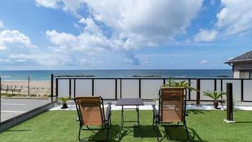 A balcony with two chairs and a table, overlooking a beach and ocean.