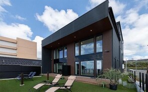 A modern building with large windows, a flat roof, and a small garden area with lounge chairs.